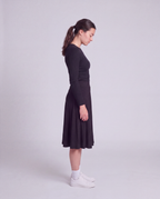 A woman in a side profile wearing a black  midi flared skirt, paired with white socks and white sneakers, standing against a light gray studio background.