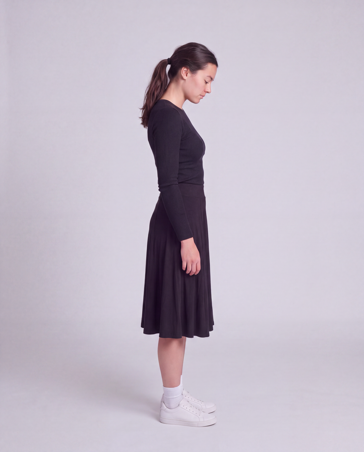 A woman in a side profile wearing a black  midi flared skirt, paired with white socks and white sneakers, standing against a light gray studio background.