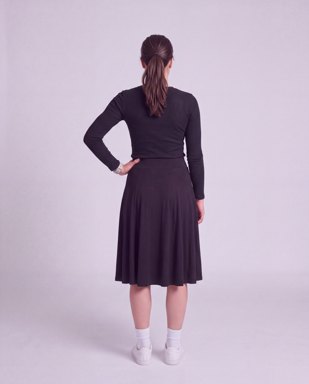 A woman is shown from behind wearing a black knee length midi skirt, paired with white socks and white sneakers, against a light gray studio background.