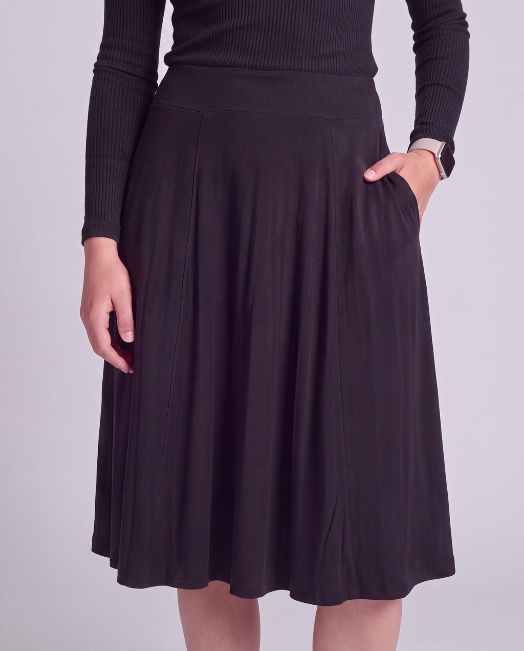 A woman wearing a black  knee length modest activwear skirt with side pockets and a matching ribbed long-sleeve top, shown from the chest down against a light gray studio background.