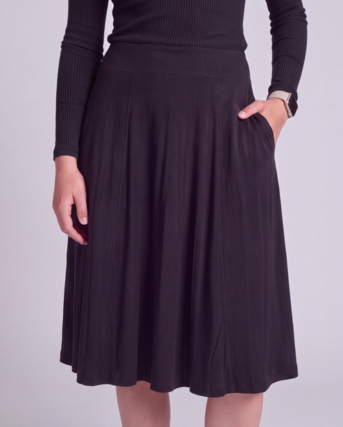 A woman wearing a black  knee length modest activwear skirt with side pockets and a matching ribbed long-sleeve top, shown from the chest down against a light gray studio background.