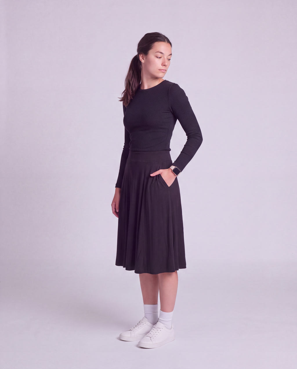 A woman modeling a modest knee length skirt side pockets, paired with white socks and white sneakers, standing against a light gray studio background.