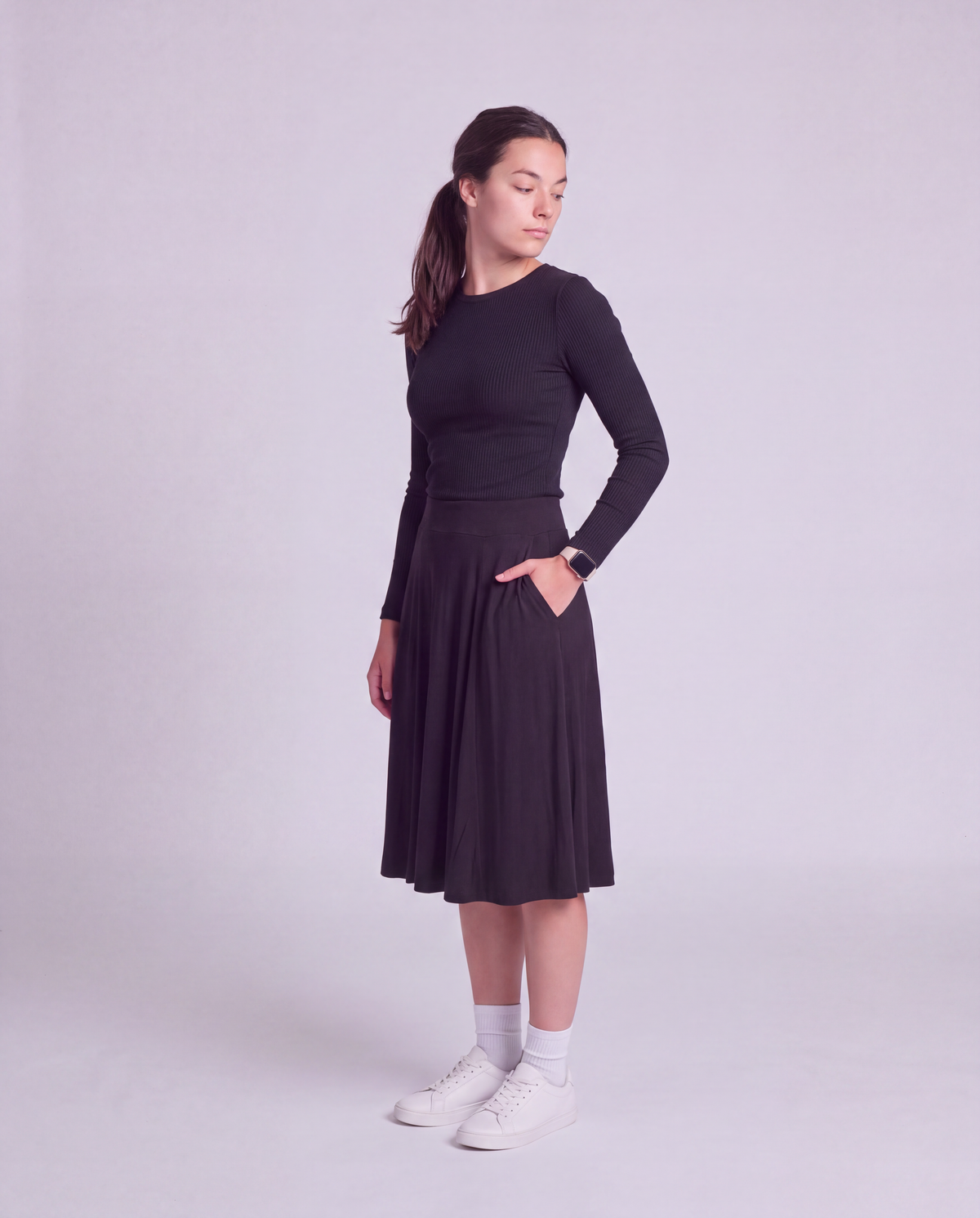 A woman modeling a modest knee length skirt side pockets, paired with white socks and white sneakers, standing against a light gray studio background.