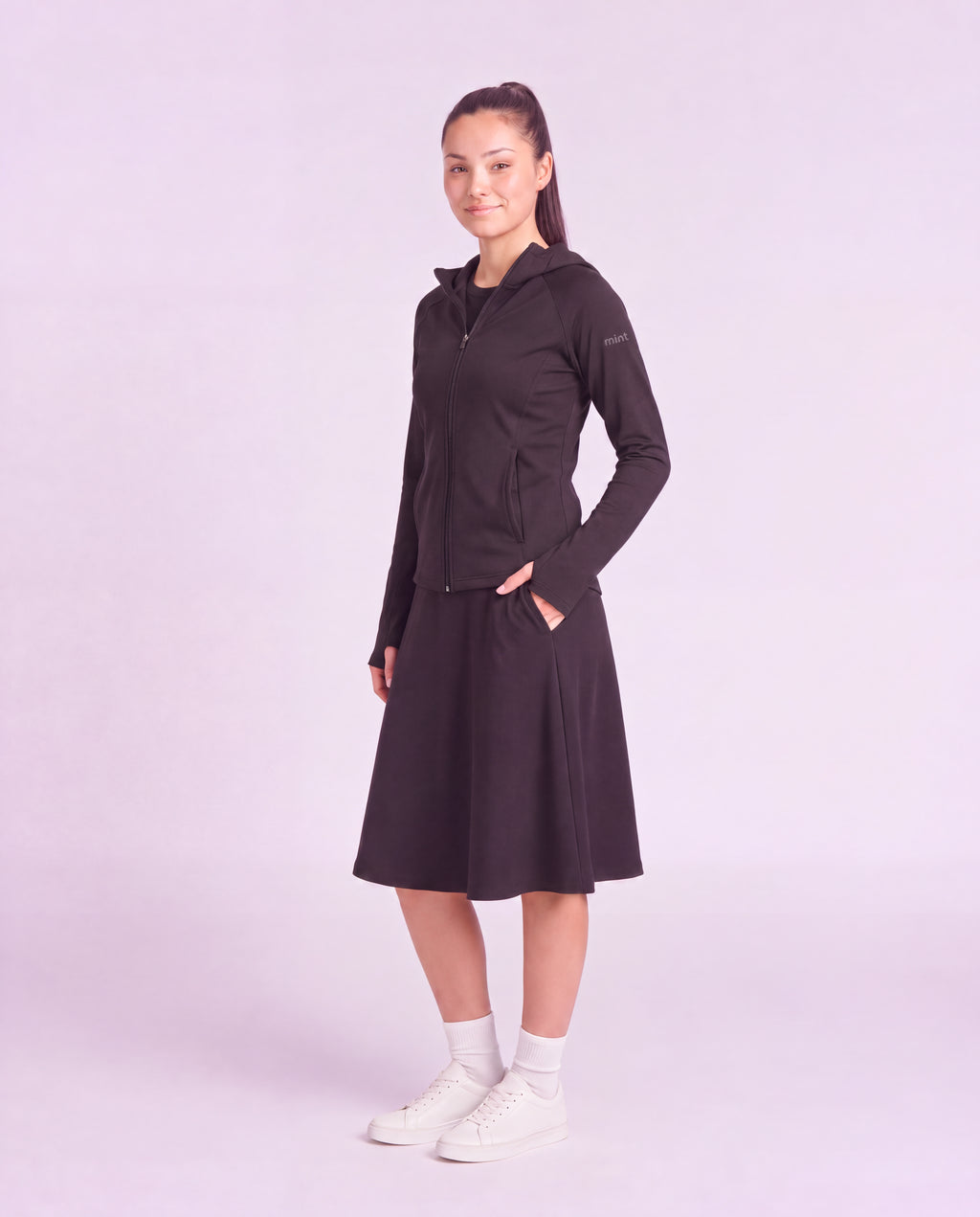 A woman modeling a black hooded zip-up jacket and matching black midi skirt set, paired with white socks and white sneakers, against a solid light lavender studio background.