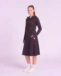 A woman modeling a black hooded zip-up jacket and matching black midi skirt set, paired with white socks and white sneakers, against a solid light lavender studio background.