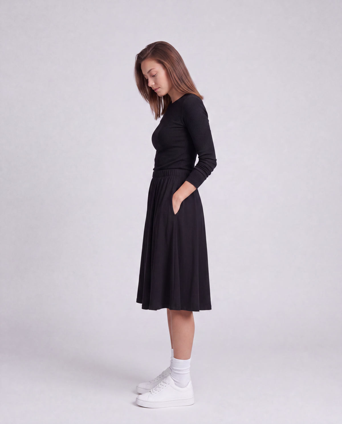 A woman modeling a black athletic modest knee length skirt with side pockets, paired with white socks and white sneakers, standing against a light gray studio background.